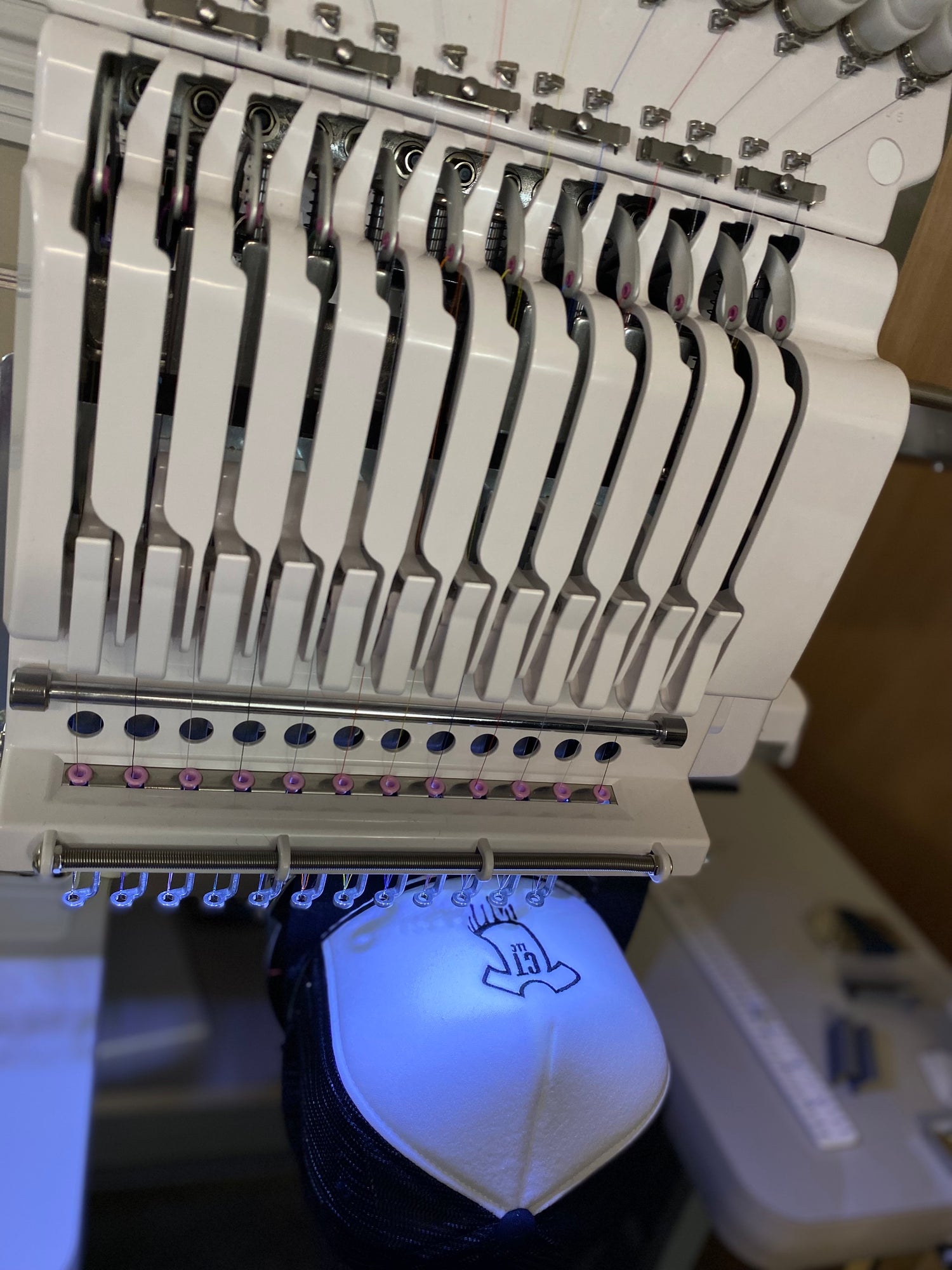 12-needle embroidery machine with white foam trucker hat with black mesh back. Embroidering Custom Threadz logo t-shirt dripping with CT LLC in the middle.